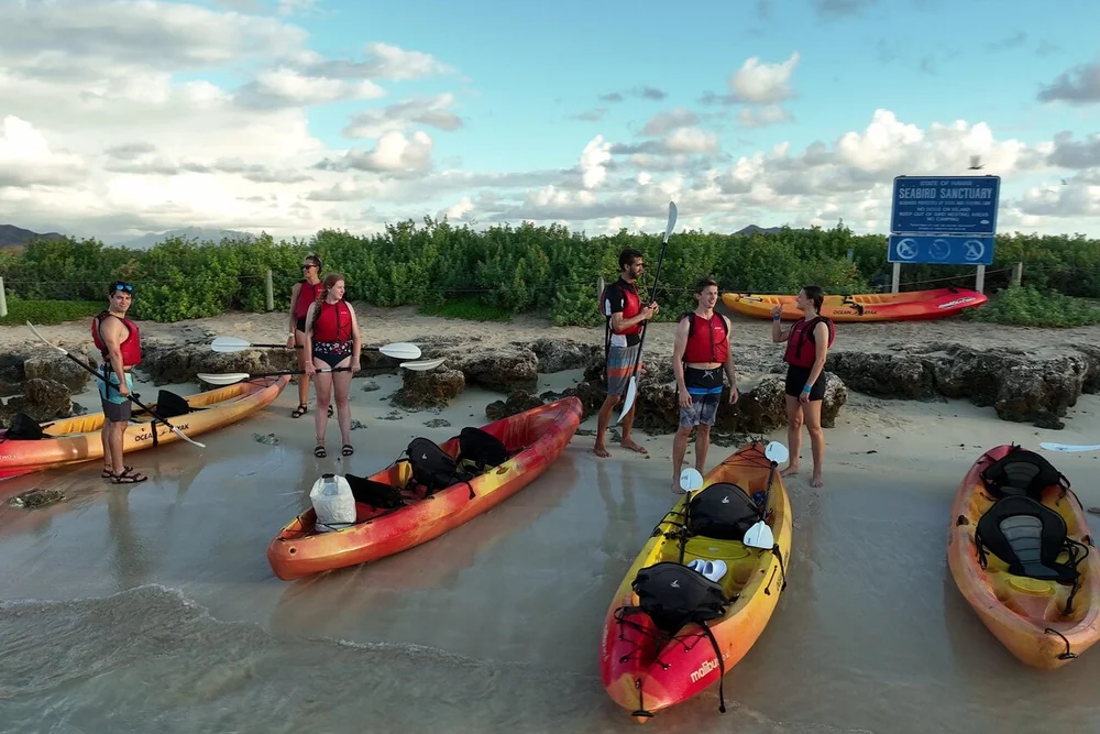 A group of kayakers in red life jackets standing with their kayaks on a small beach, a sanctuary sign behind them