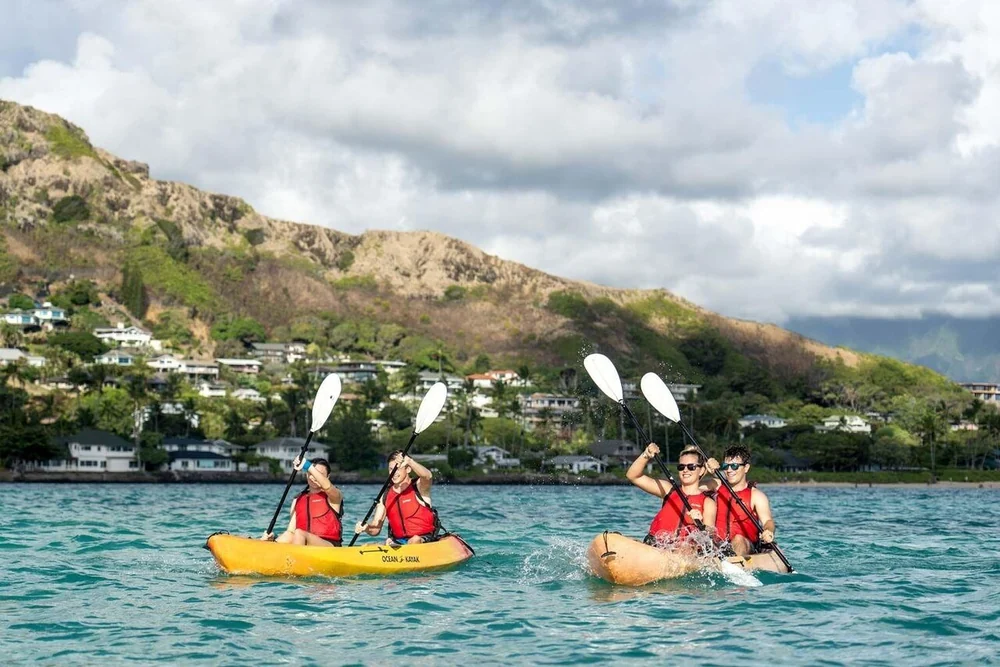 Two tandem kayaks with paddlers in red life jackets off a green hillside with houses