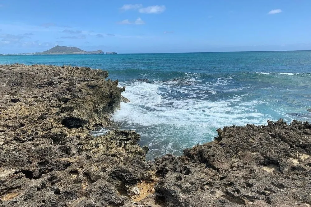 A rough, pitted rock shoreline with waves breaking into a gap, and an island headland across the water