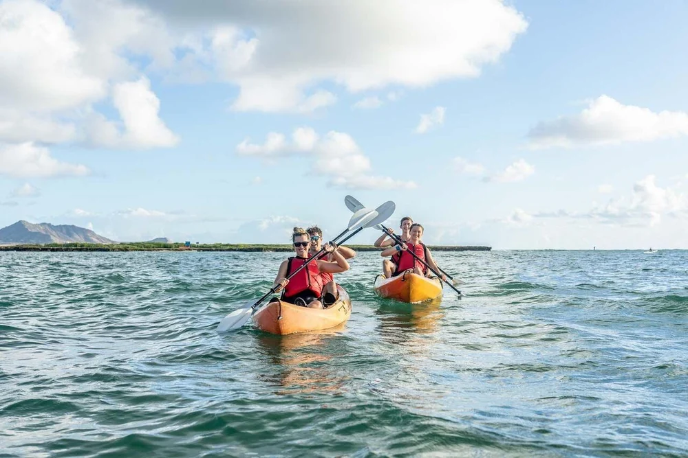 Two tandem kayaks with paddlers in red life jackets heading out across open water toward a low island